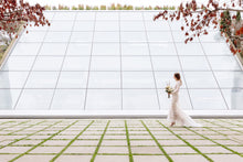 Load image into Gallery viewer, Bride walking, far back, against glass structure in Vancouver in wedding dress.