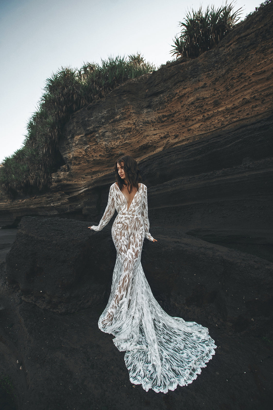 Bride leaning against black rock with long sleeve lace wedding dress fanned in front of her.