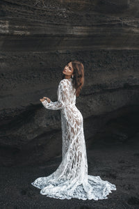 Bride with hands on rock facing away, wearing long sleeve white lace dress.