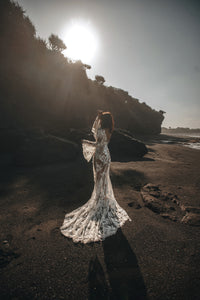 Bride on beach with hands in air showing boho sleeves and long train of wedding dress.