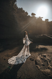 Full length side shot of model on sunny beach with long train of lace overlay wedding dress.