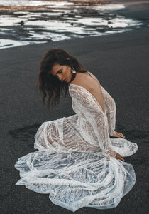 Model sitting on beach, hands in sand, showing the low back detail of lace bridal gown.