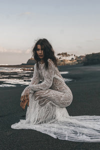 Model posing on beach with train of beaded wedding dress with long sleeve trailing behind her.